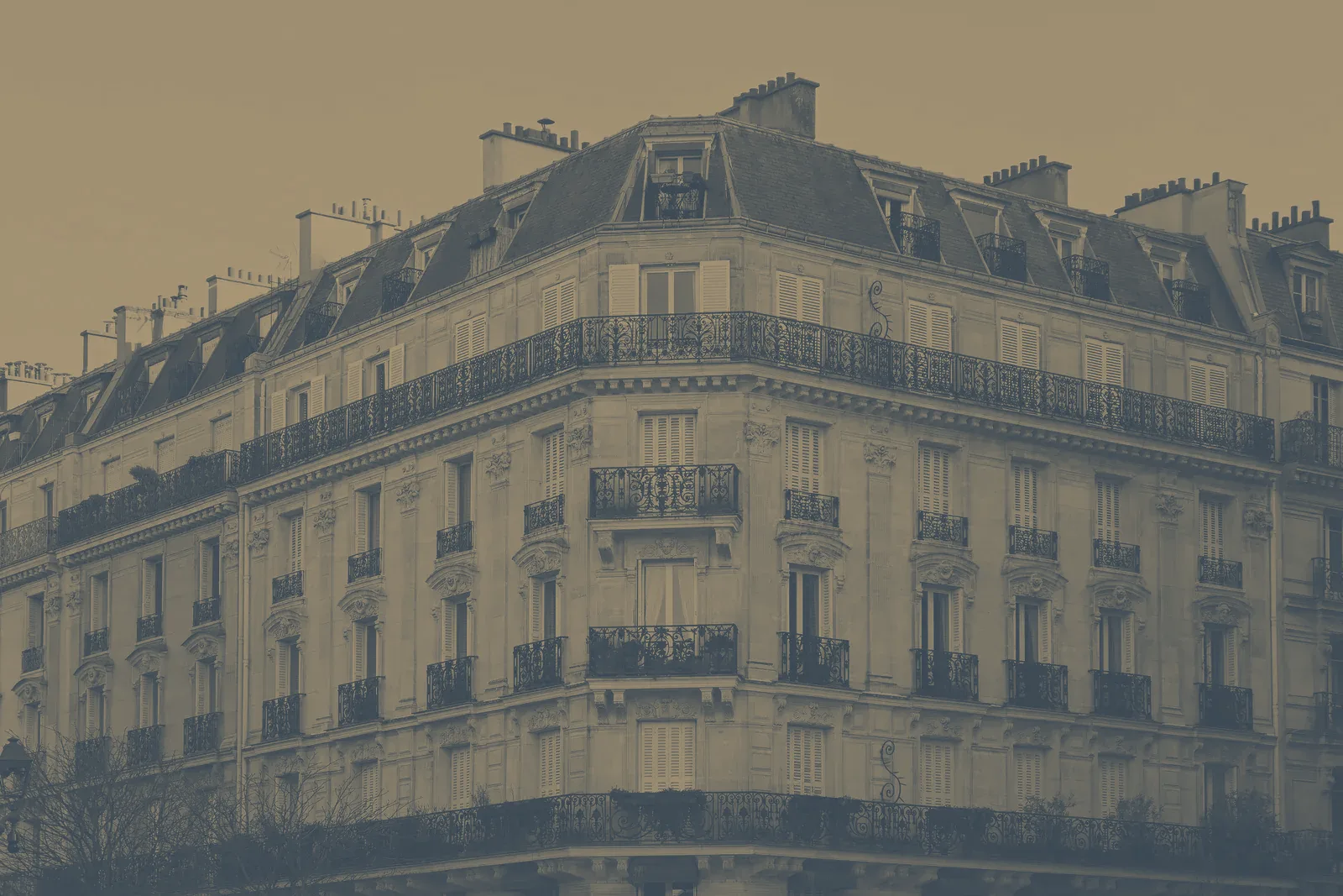 Angle d'un immeuble d'habitation haussmannien en pierre de taille, avec ses balcons en fer forgé, ses volets et son toit en ardoise, sous un ciel clair.