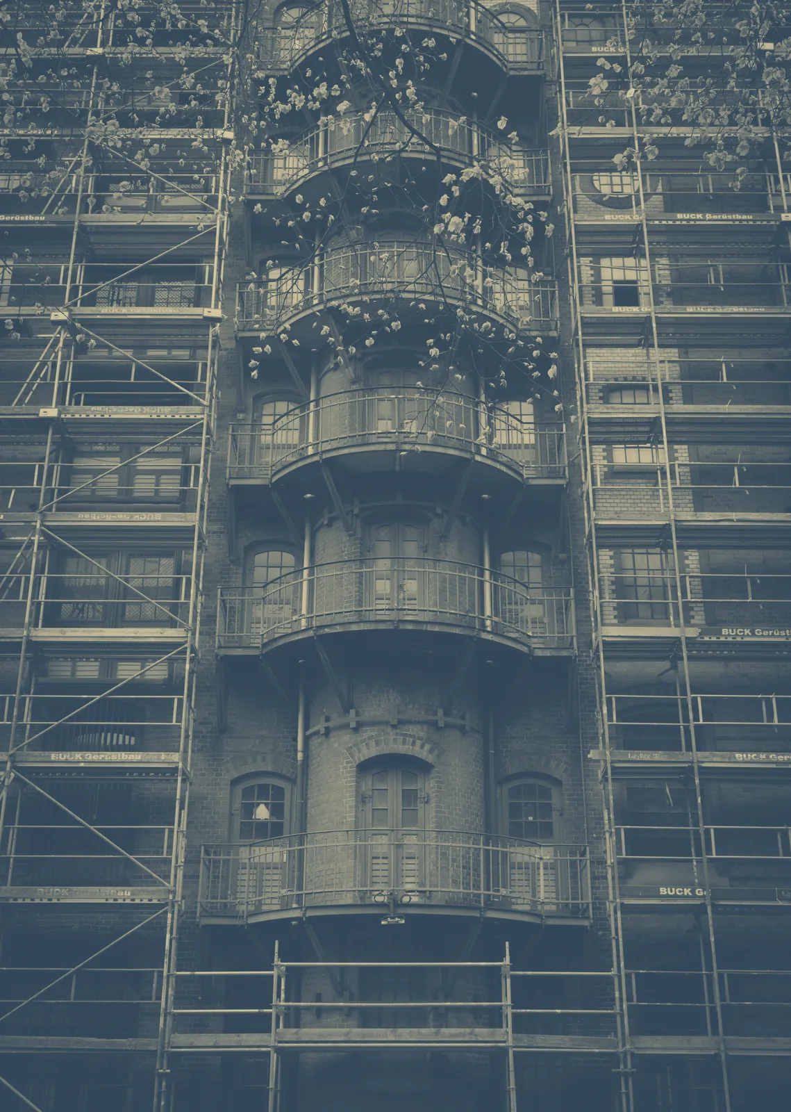 Façade d'immeuble en brique entourée d'échafaudages, avec une tour de balcons arrondis au centre et des branches en fleur au premier plan.