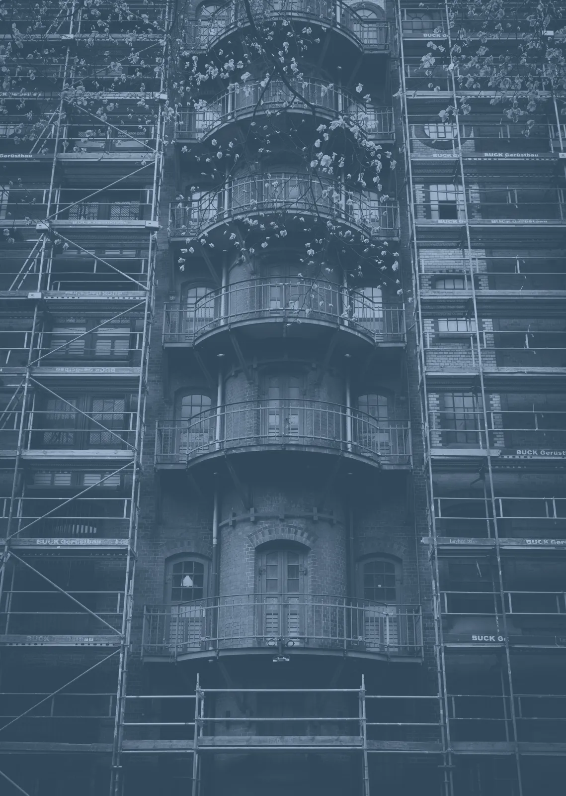 Façade d'immeuble en brique entourée d'échafaudages, avec une tour de balcons arrondis au centre et des branches en fleur au premier plan.